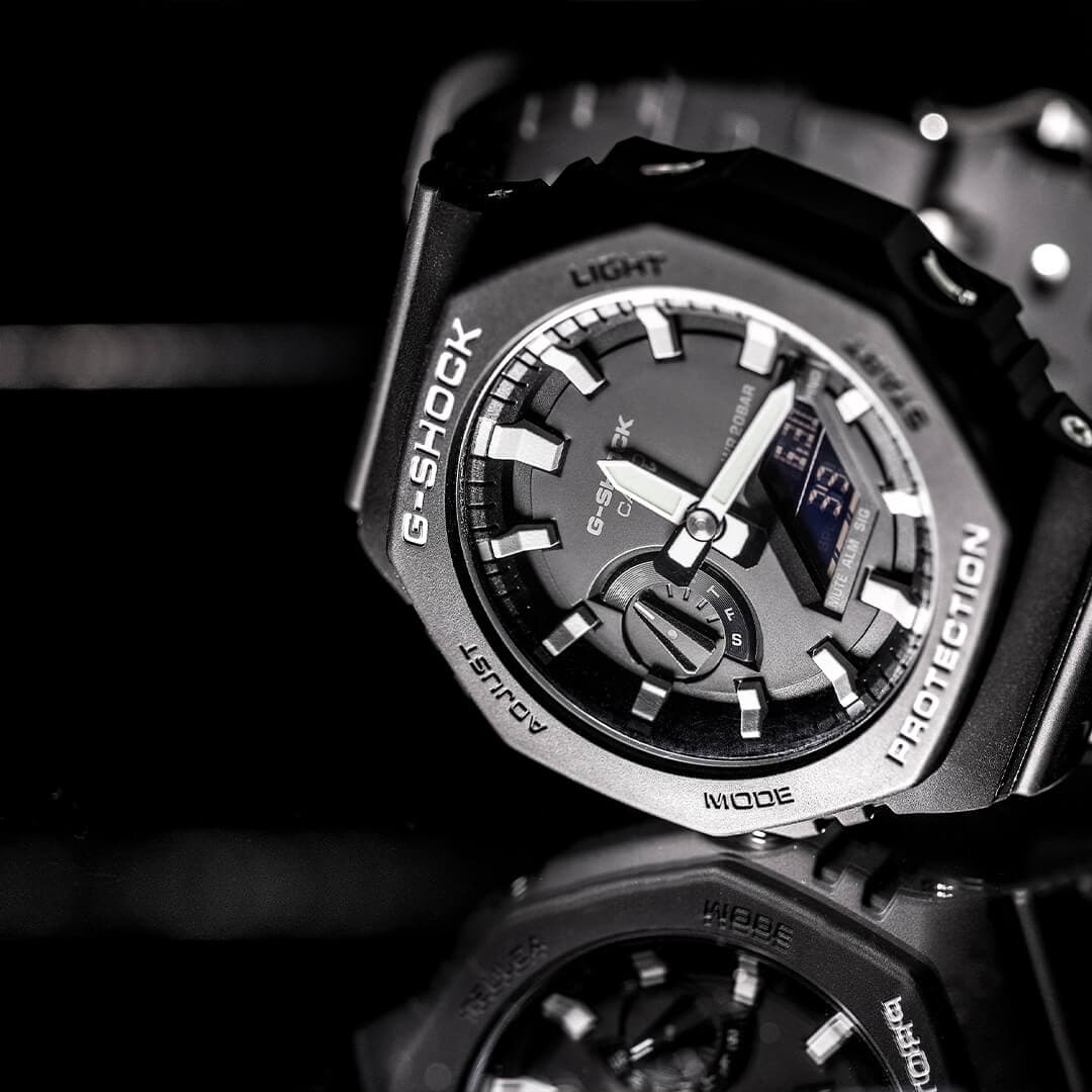 Nærbilde av en svart G-Shock-klokke med robust design, digital skjerm og metalliske detaljer, som reflekteres elegant på en blank overflate.