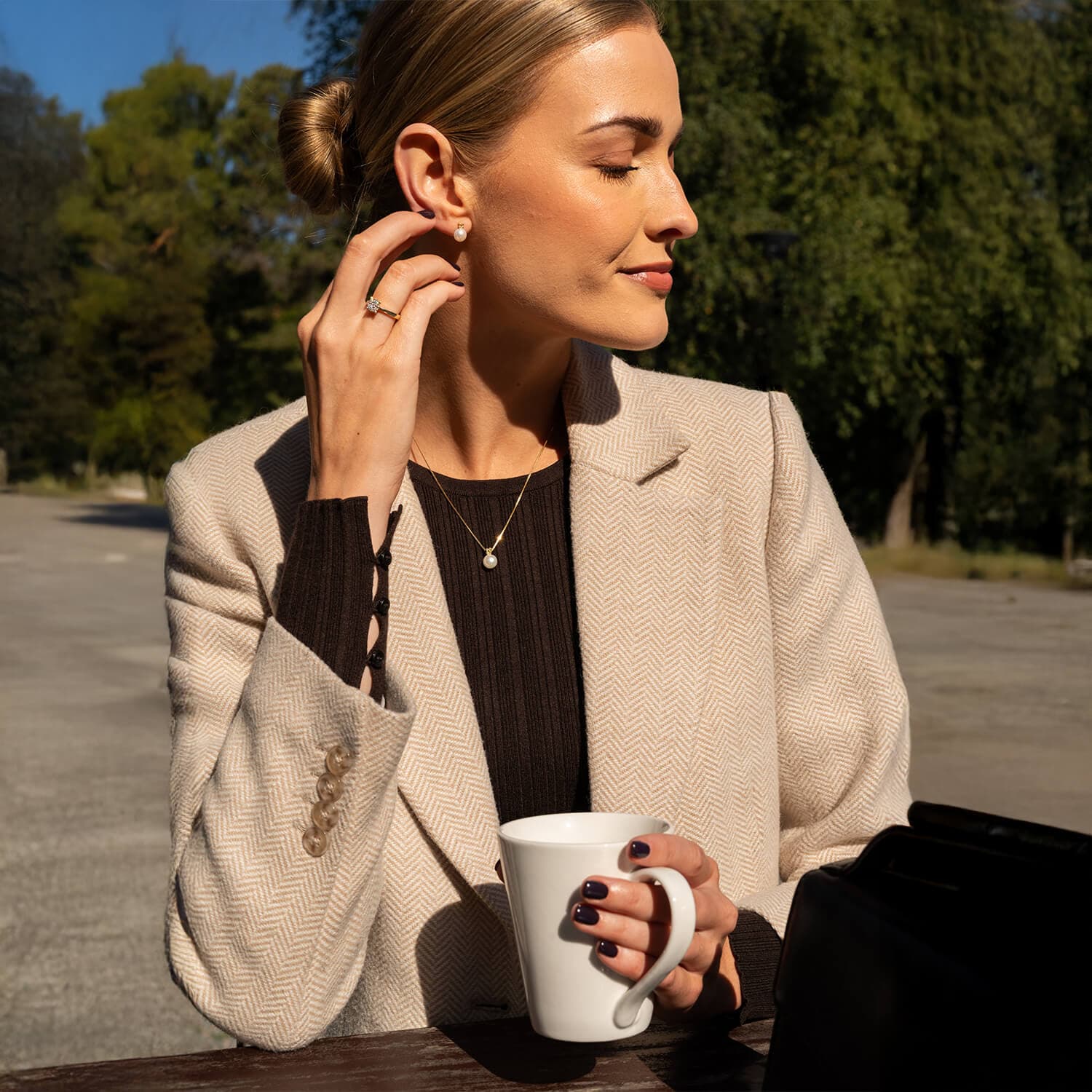 Kvinne i beige blazer koser seg med kaffe utendørs, sollys i ansiktet, trær i bakgrunnen, hånden berører øredobben hennes.