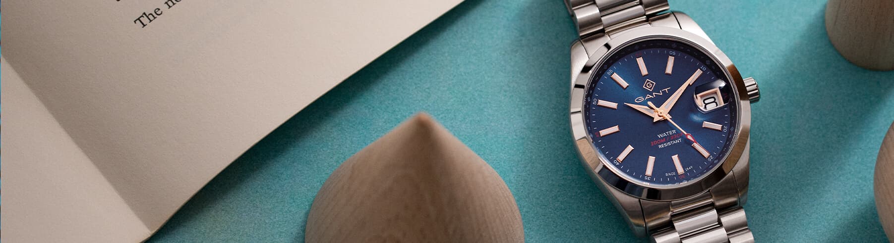 Et sølvfarget armbåndsur med en blå urskive som viser datoen står på en blågrønn overflate i nærheten av en åpen bok og tregjenstander.