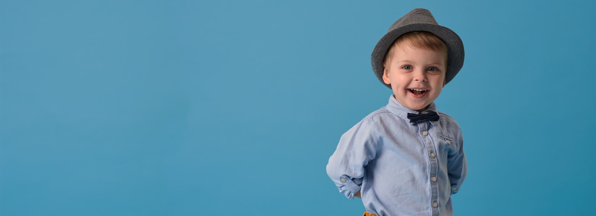 En ung gutt i blå skjorte og hatt smiler med hendene bak ryggen mot en blå bakgrunn.