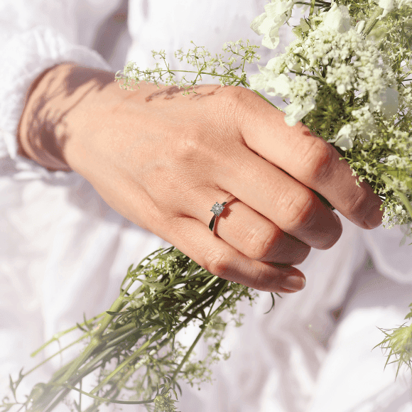 En hånd med en diamantring holder en bukett delikate hvite blomster mot en lys bakgrunn.