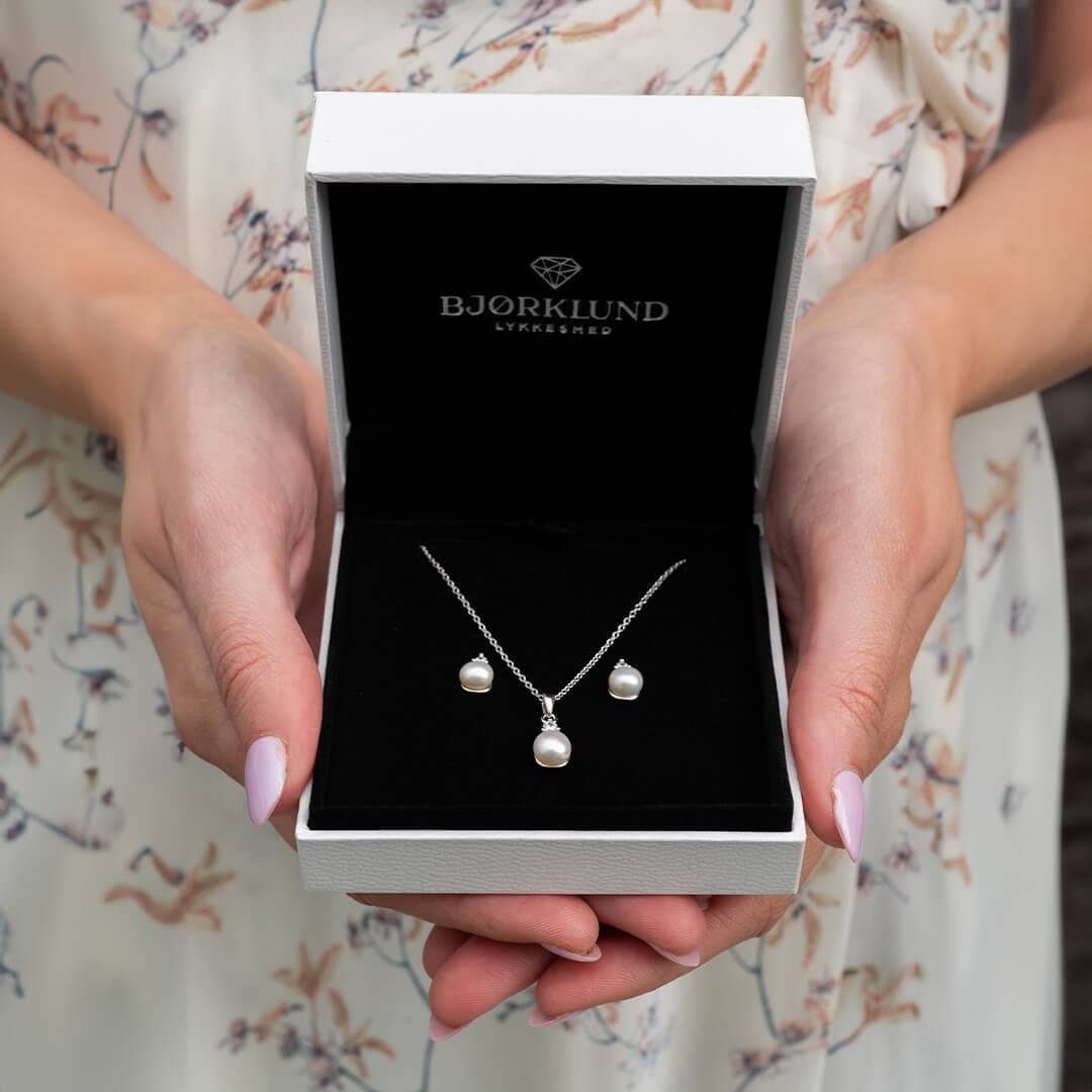 Hender holder et åpent smykkeskrin med perlekjede og matchende øredobber mot en bakgrunn av en blomstret kjole.