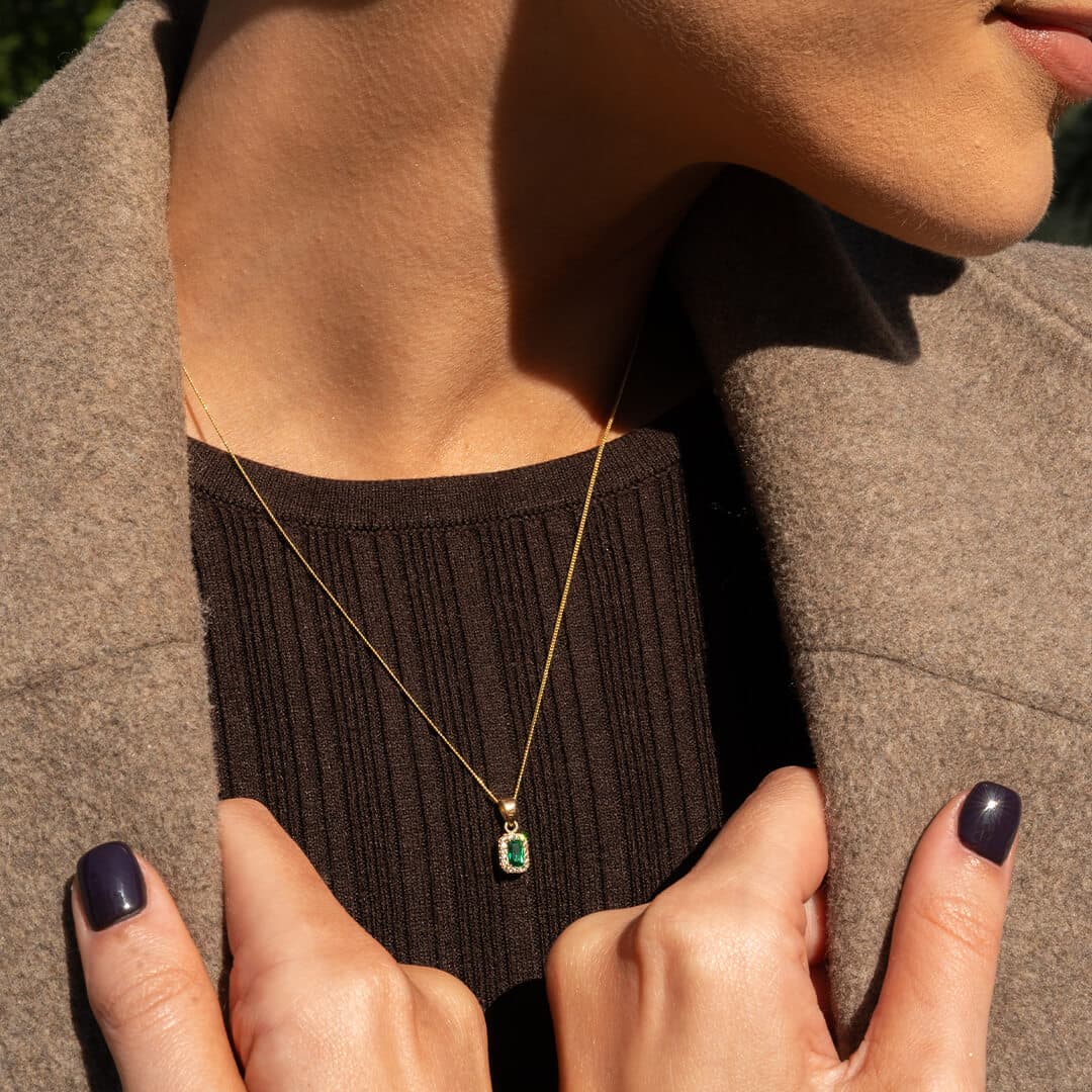 Kvinne med gullkjede med grønt anheng, brun genser og beige kåpe. Negler lakkert mørk lilla.