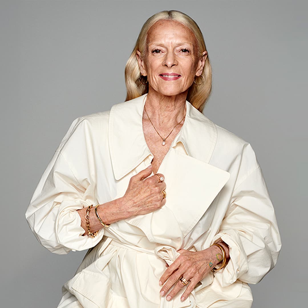 Eldre kvinne med langt hvitt hår i kremhvit kåpe. Hun poserer mot grå bakgrunn med et rolig blikk og ringer på hånden.