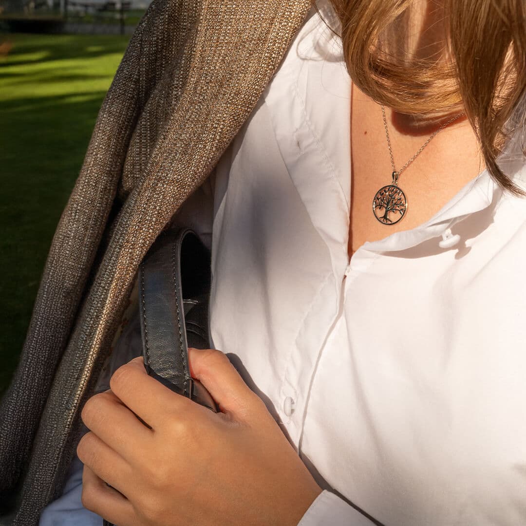 Person iført en brun frakk drapert over skuldrene, hvit skjorte og et halskjede med treanheng, som holder en grå håndveskestropp.