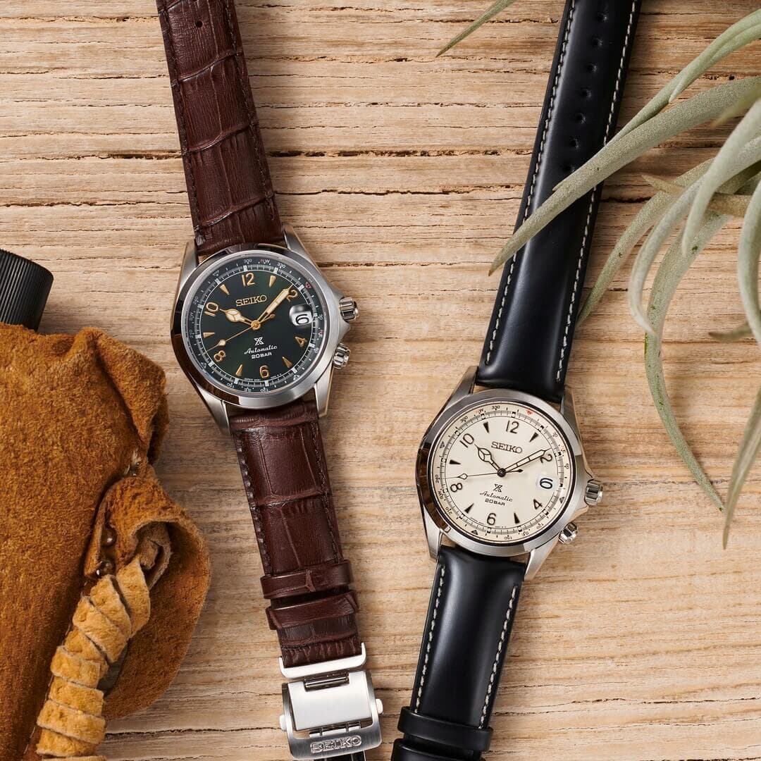 To Seiko-klokker på en treoverflate. Den ene har en brun lærreim og svart urskive, den andre en svart lærreim og kremfarget urskive.