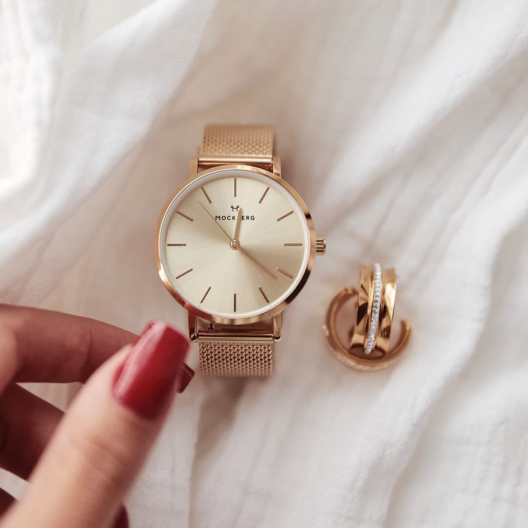 Et gullfarget armbåndsur med nettingrem og tre gullringer på en hvit stoffbakgrunn. En hånd med røde negler justerer klokken.