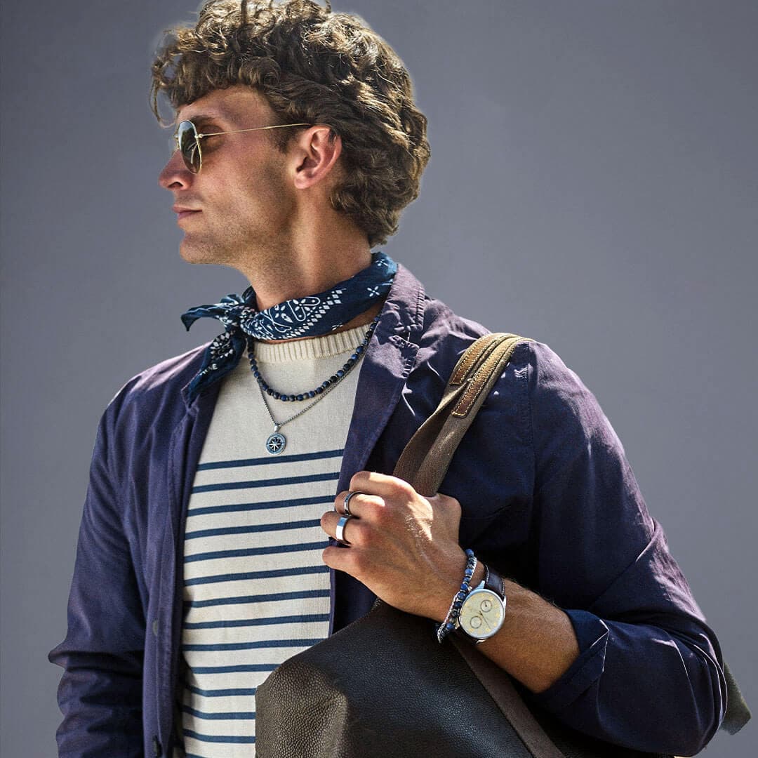 Mann med solbriller og stripete skjorte, iført blå jakke, bandana og halskjeder, holder en skinnveske på skulderen mot grå bakgrunn.