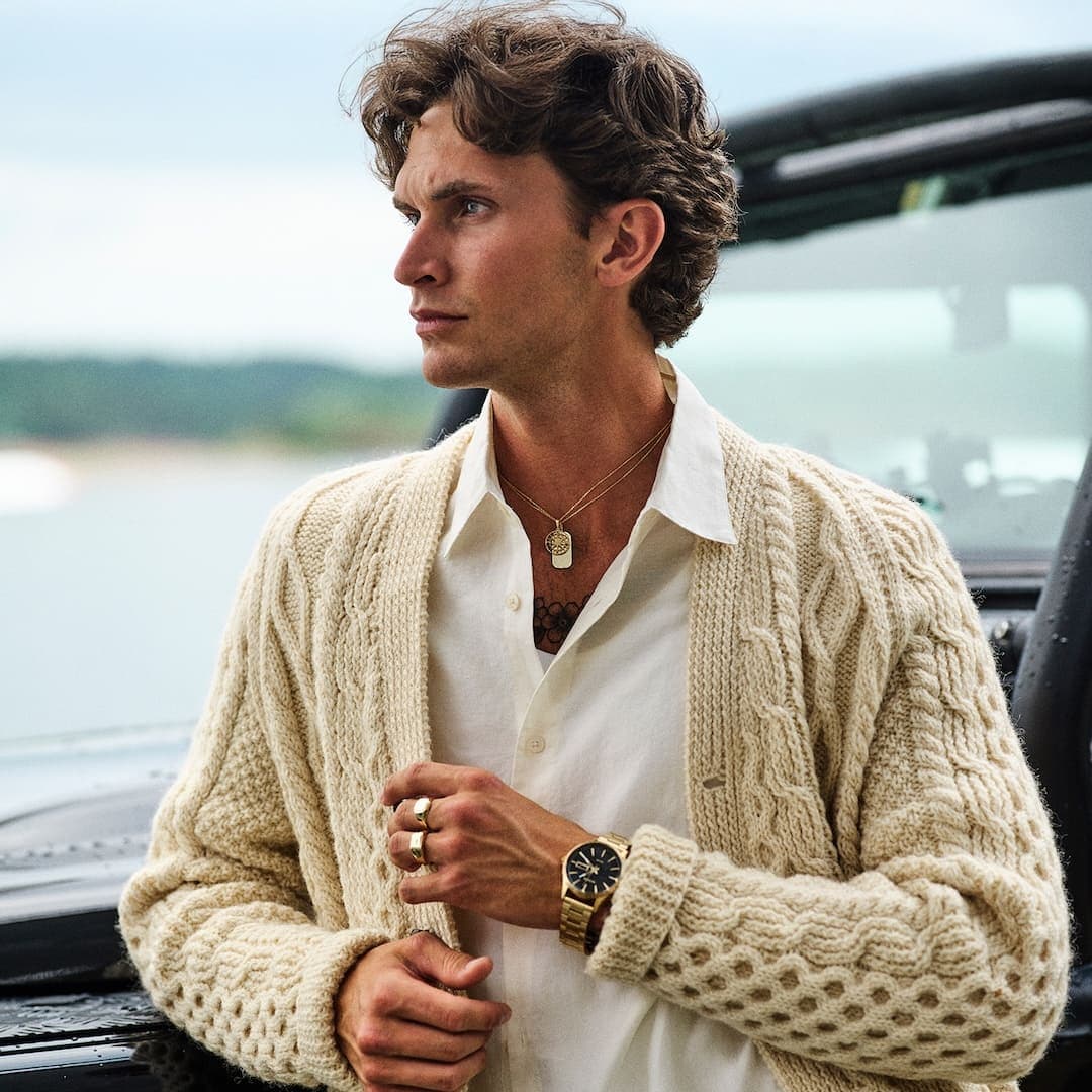 Mann i hvit skjorte og kremfarget cardigan, med smykker og klokke, står ved et kjøretøy med vann og trær i bakgrunnen.