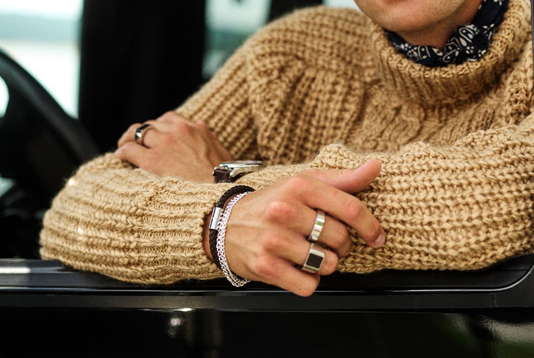 Nærbilde av hendene til en stående mann med beige, tynn ullgenser, som holder på gullringen som er på tommelen hans..