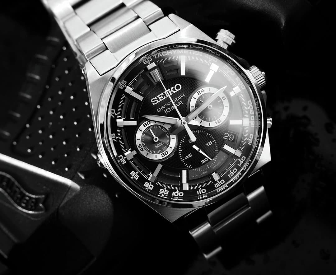 Seiko kronograf med stållenke, sort urskive, flere underurskiver og takymeterskala vist i nærbilde. Stilrent design.