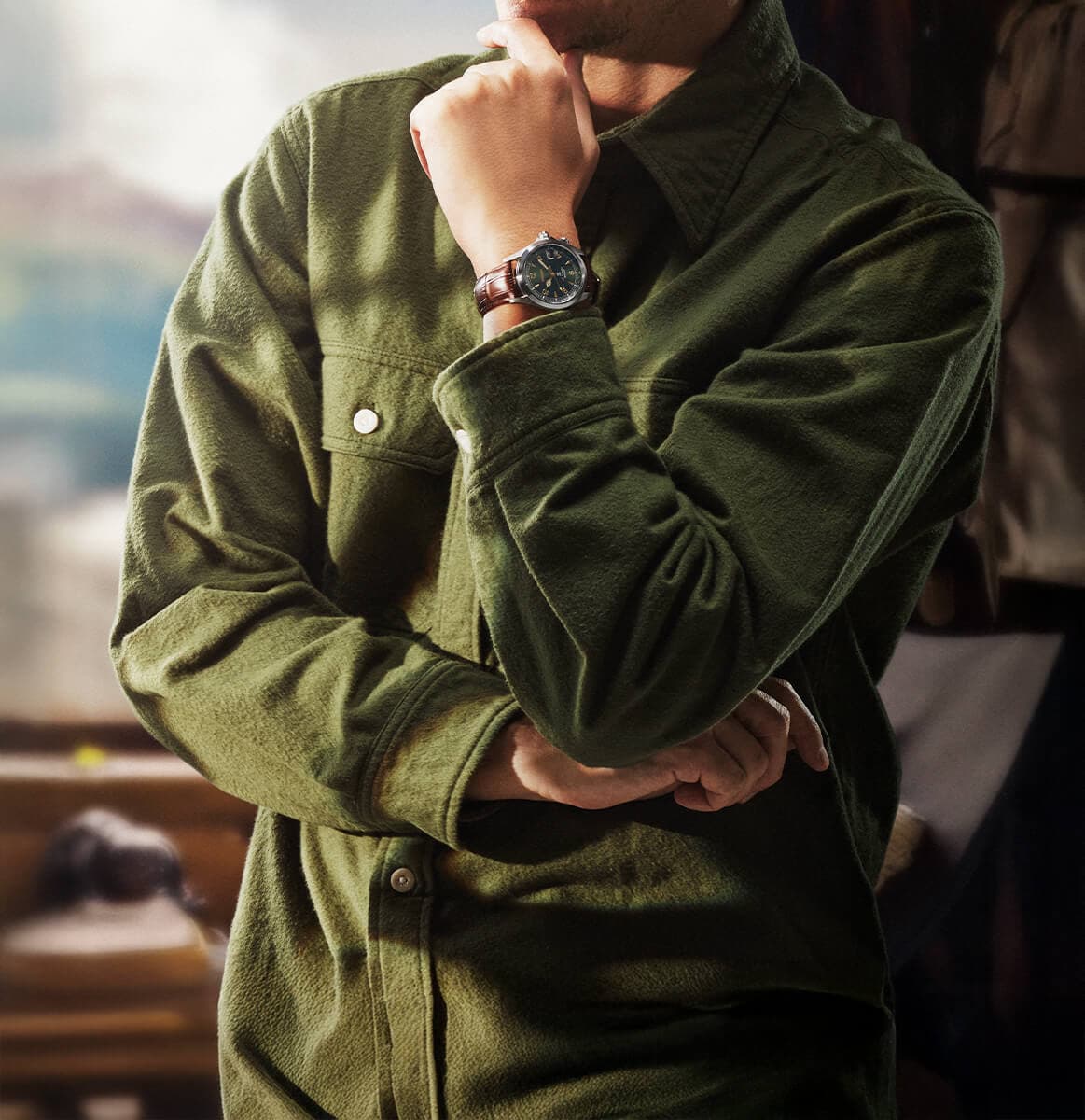 Mann i grønn skjorte poserende innendørs med én hånd på haken og den andre armen i kors, iført et armbåndsur.