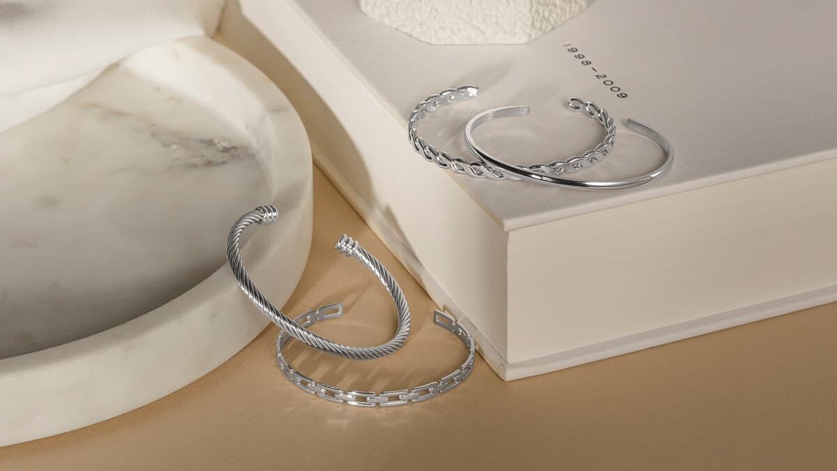 Sølvarmbånd er utstilt på en beige overflate, med en åpen bok og et marmorfat i bakgrunnen.