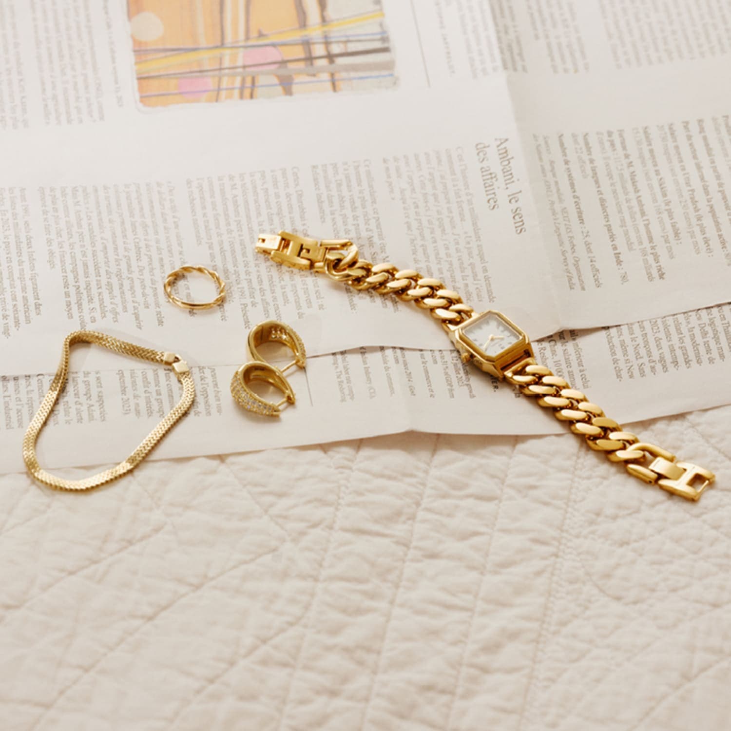 Gullklokke, armbånd og ringer presentert på en avis, plassert på en vattert overflate. Minimalistisk og elegant oppstilling.