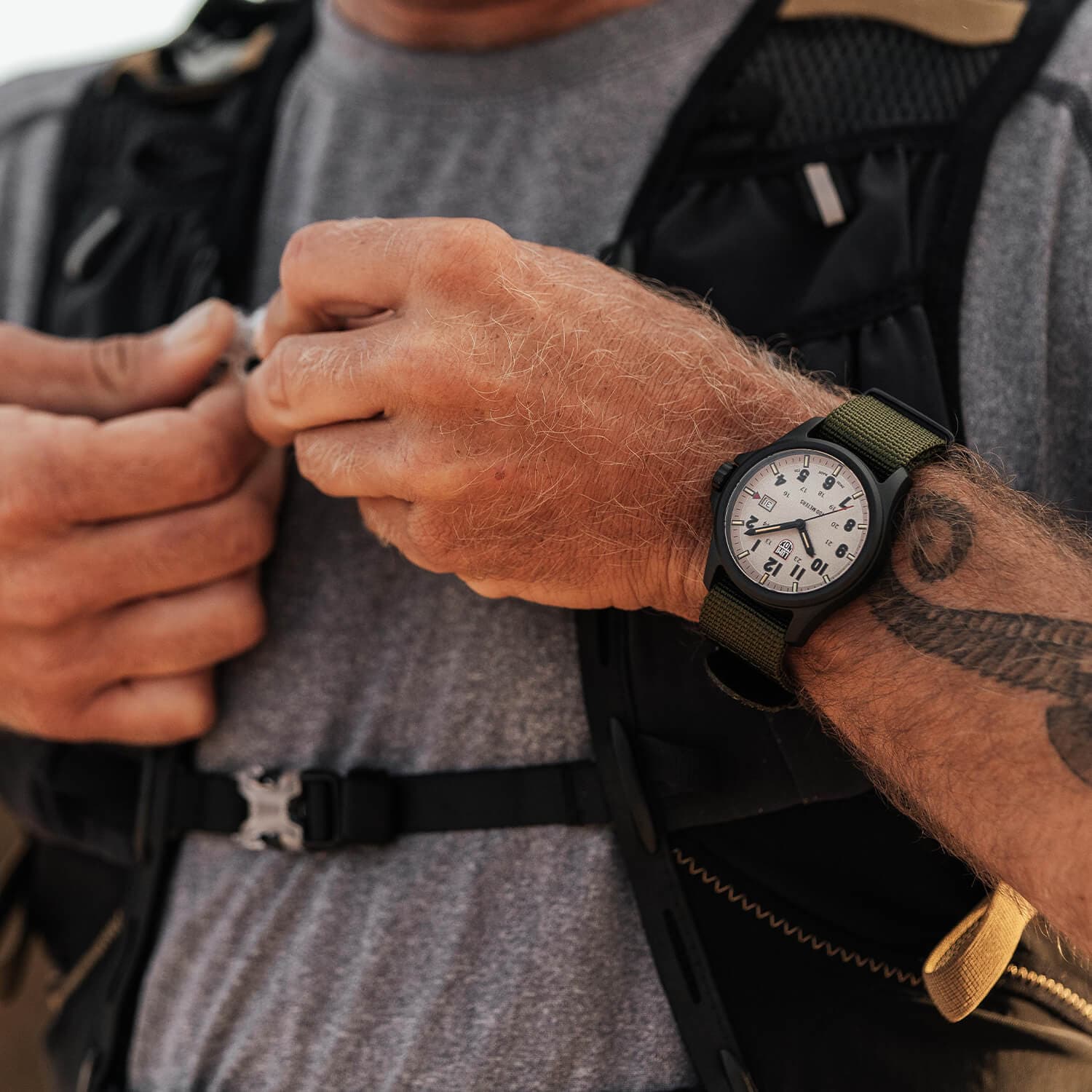 Person som retter på en svart taktisk vest, iført en grønn armbåndsur med hvit urskive og en synlig tatovering på underarmen.