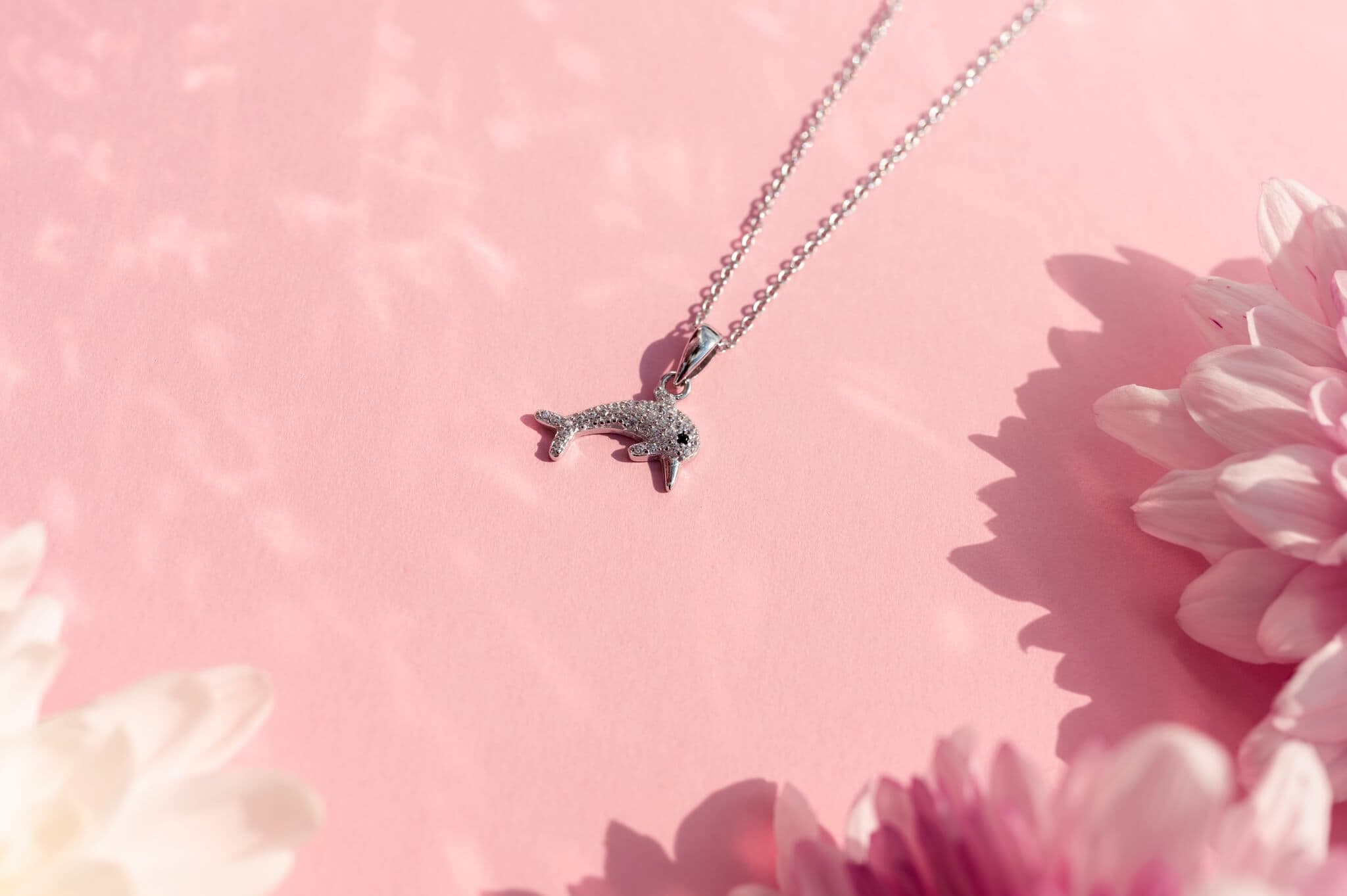Sølvfarget delfinhalskjede på rosa bakgrunn, omgitt av myke rosa og hvite blomster.
